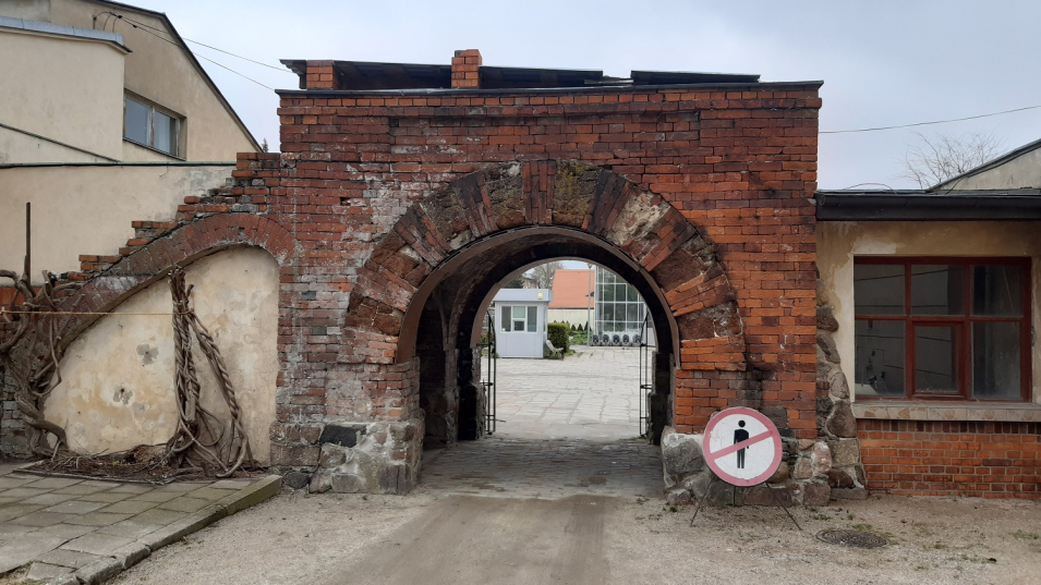 Ceglana brama z półkolistym przejściem. Po bokach szaro-beżowe zabudowania