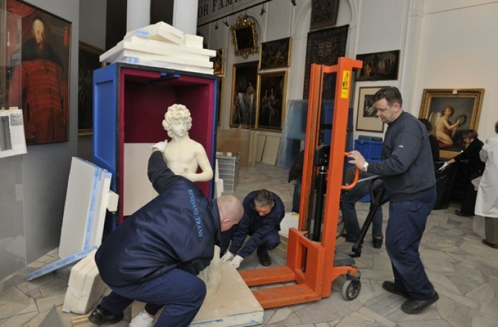 Pracownicy zajmują się zabezpieczaniem i przenoszeniem rzeźby w muzeum. Posąg umieszczany jest w specjalnym pojemniku wyłożonym ochronnym materiałem. Obok znajduje się podnośnik, który pomaga w transporcie. W tle widoczne są obrazy na ścianach.