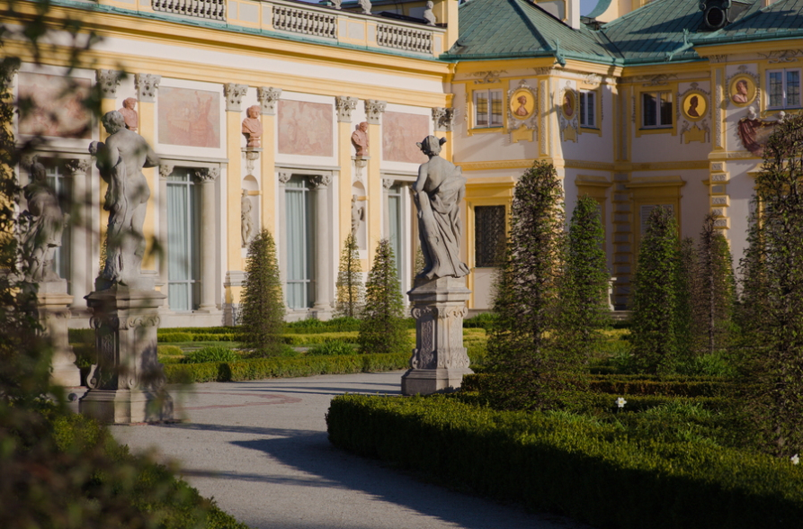 Widok na fragment pałacu w Wilanowie od strony ogrodu. Wśród roślinności stoi rzeźba na cokole.