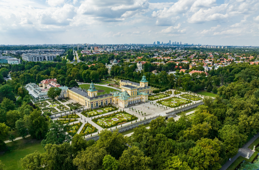 Pałac w Wilanowie widziany z lotu ptaka. Otoczony się drzewami. Zdjęcie zostało zrobione w porze letniej. W tle zabudowania Warszawy.